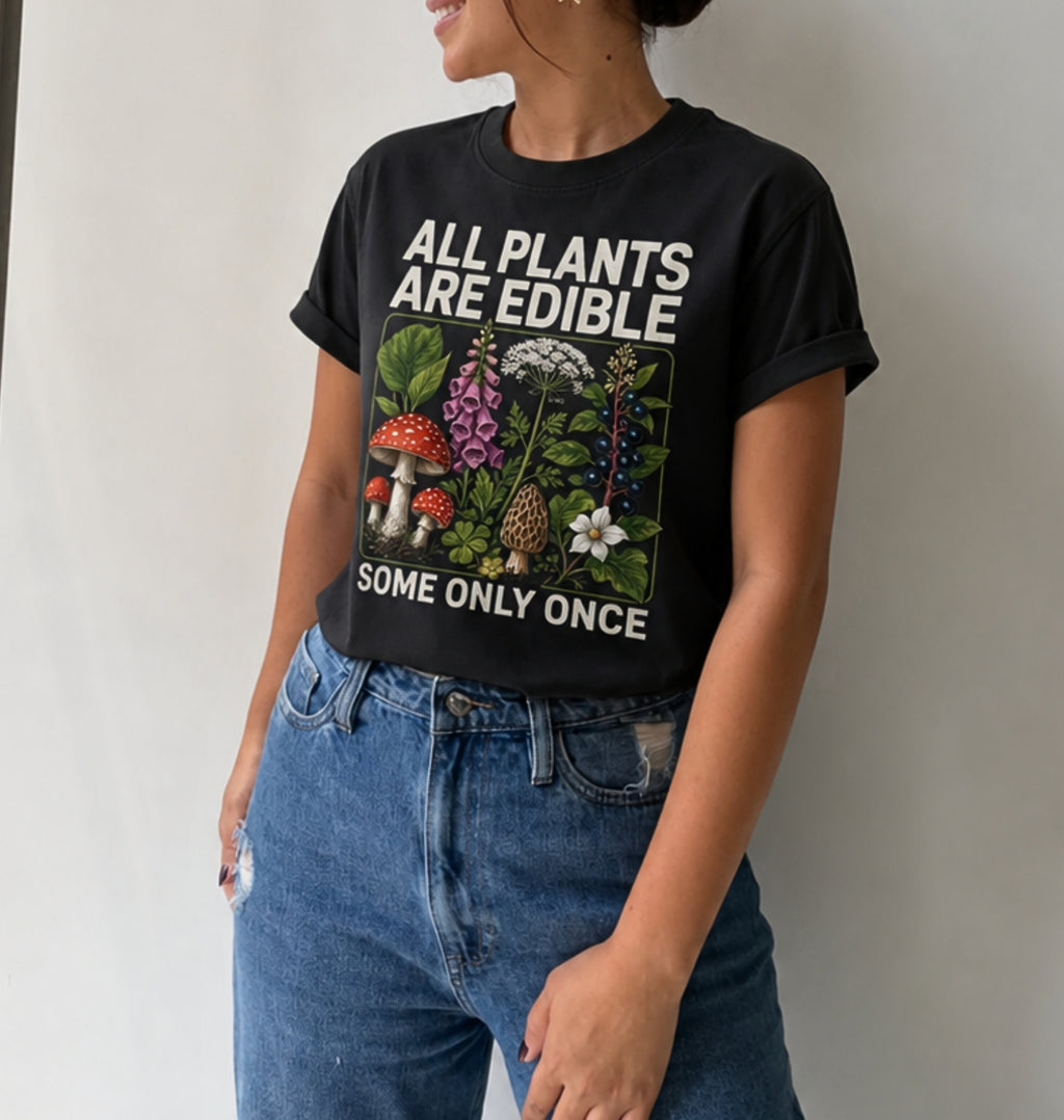 A woman wearing a black graphic t-shirt with the text 'ALL PLANTS ARE EDIBLE SOME ONLY ONCE' and a colorful illustration of edible plants and mushrooms, paired with blue jeans.