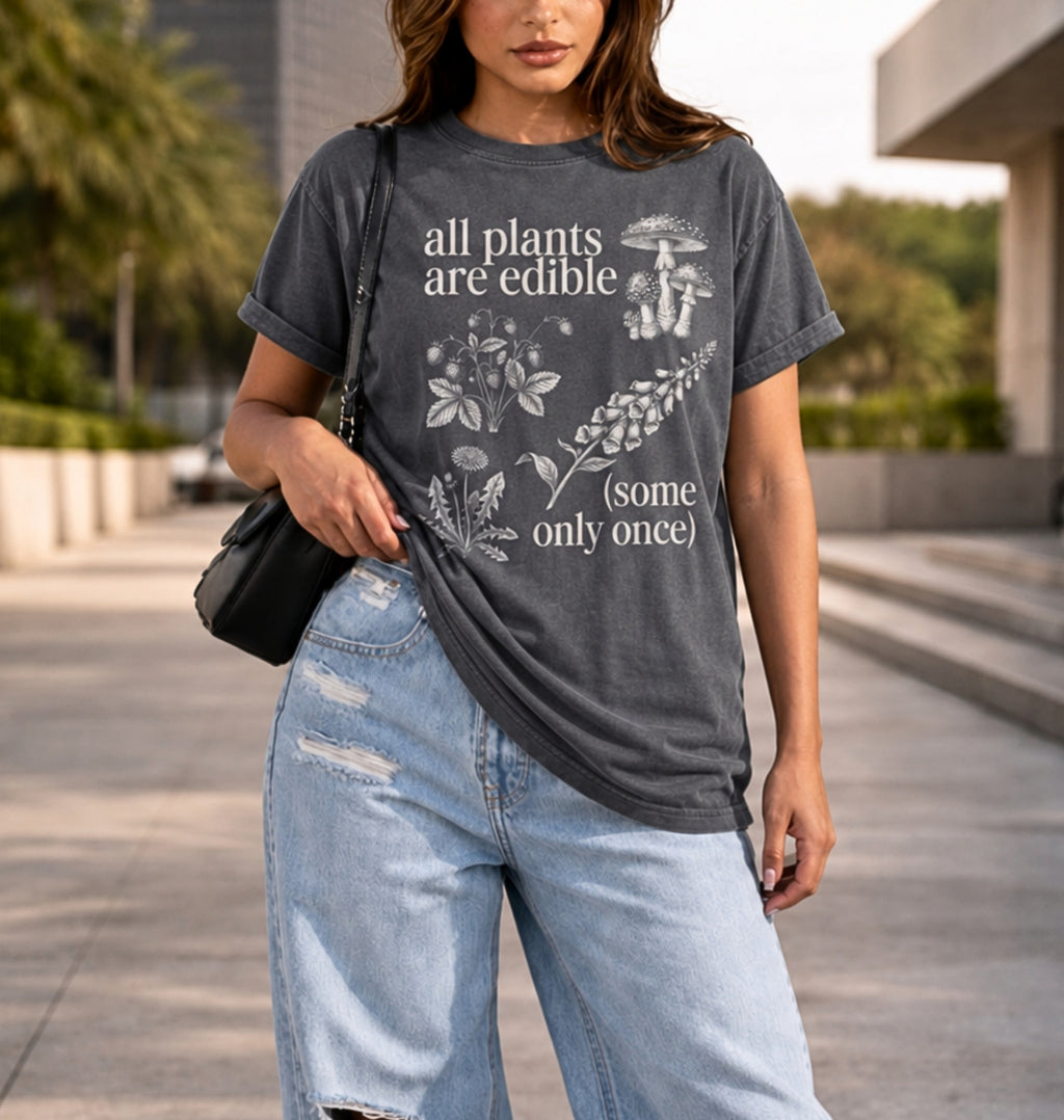 A woman wearing a dark gray graphic t-shirt with white text reading 'all plants are edible (some only once)' and botanical illustrations, paired with light-wash distressed jeans and a black shoulder bag, standing outdoors on a paved walkway.