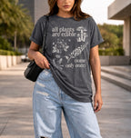 A woman wearing a dark gray graphic t-shirt with white text reading 'all plants are edible (some only once)' and botanical illustrations, paired with light-wash distressed jeans and a black shoulder bag, standing outdoors on a paved walkway.