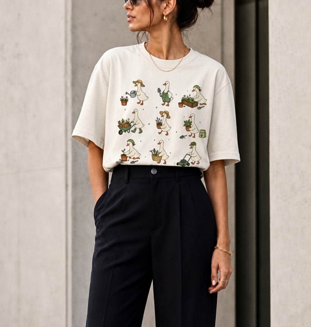 A woman wearing a cream-colored short-sleeve t-shirt with a vintage-style cartoon duck print, paired with black high-waisted trousers, standing against a neutral concrete wall.