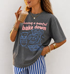 A woman wearing a dark gray graphic t-shirt with the text 'having a mental bake down' in pink and blue doodle illustrations, paired with blue and white striped pants, holding an iced coffee.