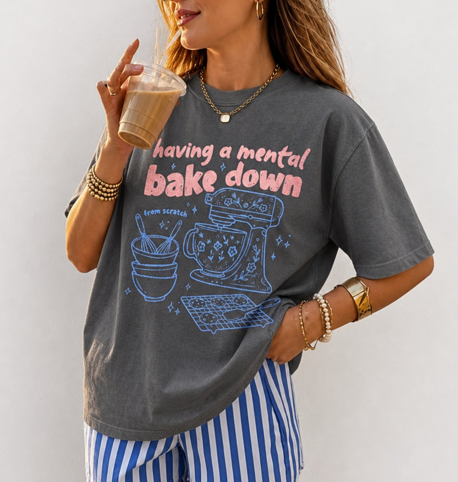 A woman wearing a dark gray graphic t-shirt with the text 'having a mental bake down' in pink and blue doodle illustrations, paired with blue and white striped pants, holding an iced coffee.
