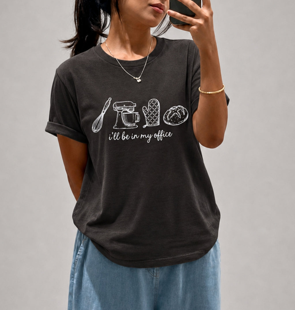 A woman takes a mirror selfie wearing a dark charcoal gray t-shirt with white line drawings of kitchen utensils and the phrase 'I'll be in my office'. She is wearing light blue jeans and a gold necklace.