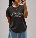 A woman takes a mirror selfie wearing a dark charcoal gray t-shirt with white line drawings of kitchen utensils and the phrase 'I'll be in my office'. She is wearing light blue jeans and a gold necklace.