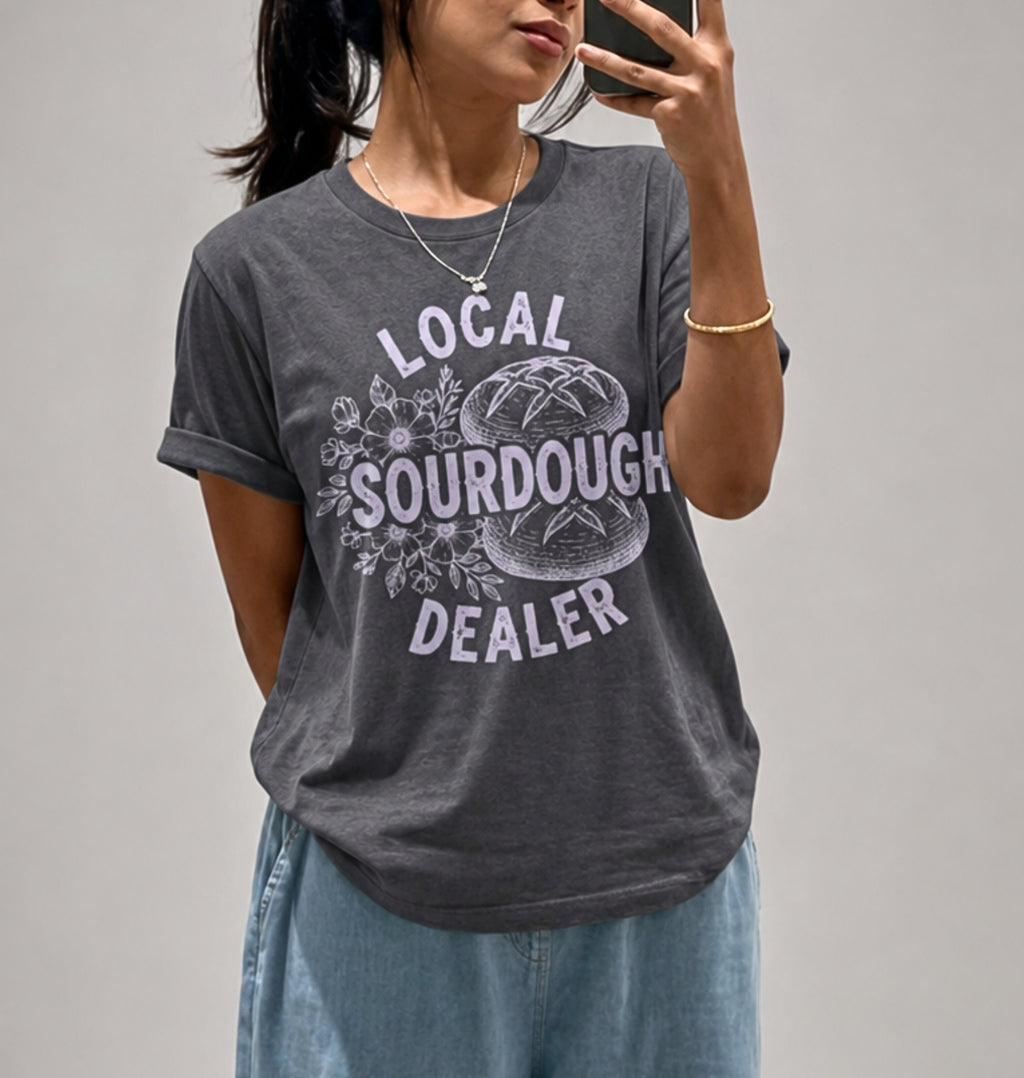 A woman takes a mirror selfie wearing a dark gray graphic t-shirt with a white distressed print that reads 'LOCAL SOURDOUGH DEALER' around a sourdough bread illustration. She is also wearing light blue jeans and a gold necklace.