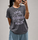 A woman takes a mirror selfie wearing a dark gray graphic t-shirt with a white distressed print that reads 'LOCAL SOURDOUGH DEALER' around a sourdough bread illustration. She is also wearing light blue jeans and a gold necklace.