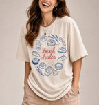 A woman wearing a cream-colored graphic t-shirt with a blue and brown illustration of baking ingredients and utensils, standing in a kitchen setting.