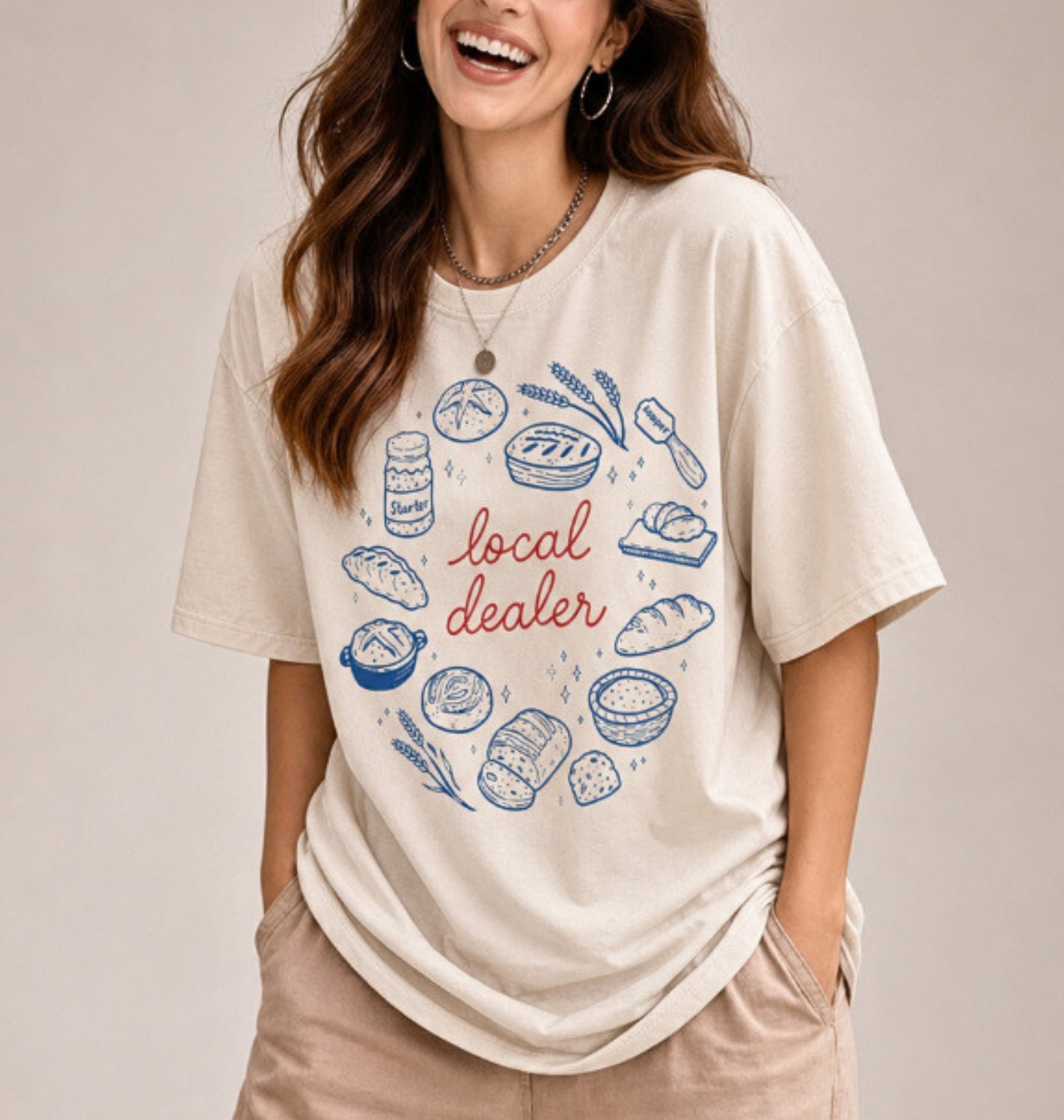 A woman wearing a cream-colored graphic t-shirt with a blue and brown illustration of baking ingredients and utensils, standing in a kitchen setting.