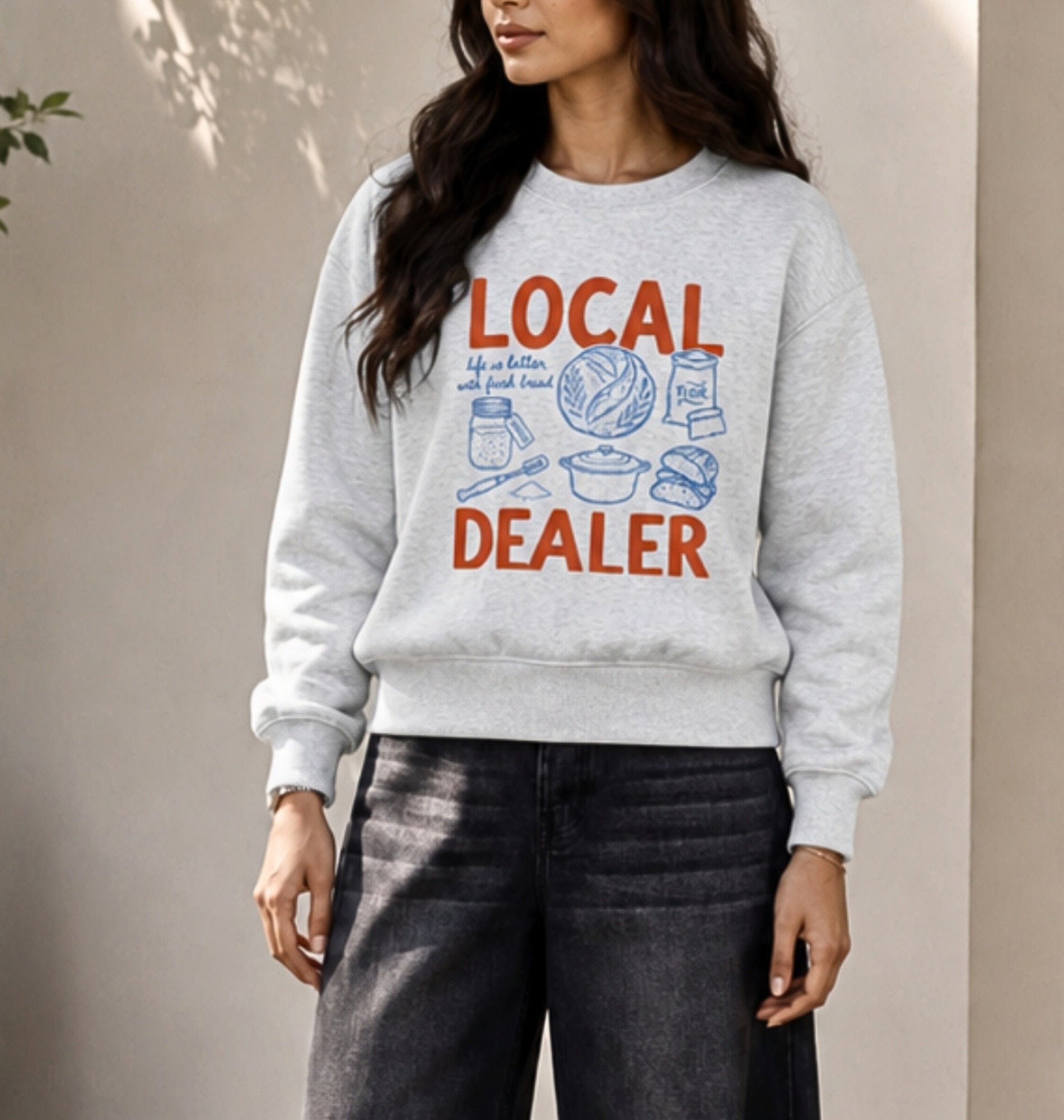 A woman wearing a light heather gray crewneck sweatshirt with a retro-style graphic that reads 'LOCAL DEALER' in orange-red and blue, paired with dark gray jeans.