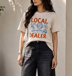 A woman wearing a beige graphic t-shirt with orange and blue text reading 'LOCAL DEALER' and dark gray wide-leg jeans, standing against a neutral wall.