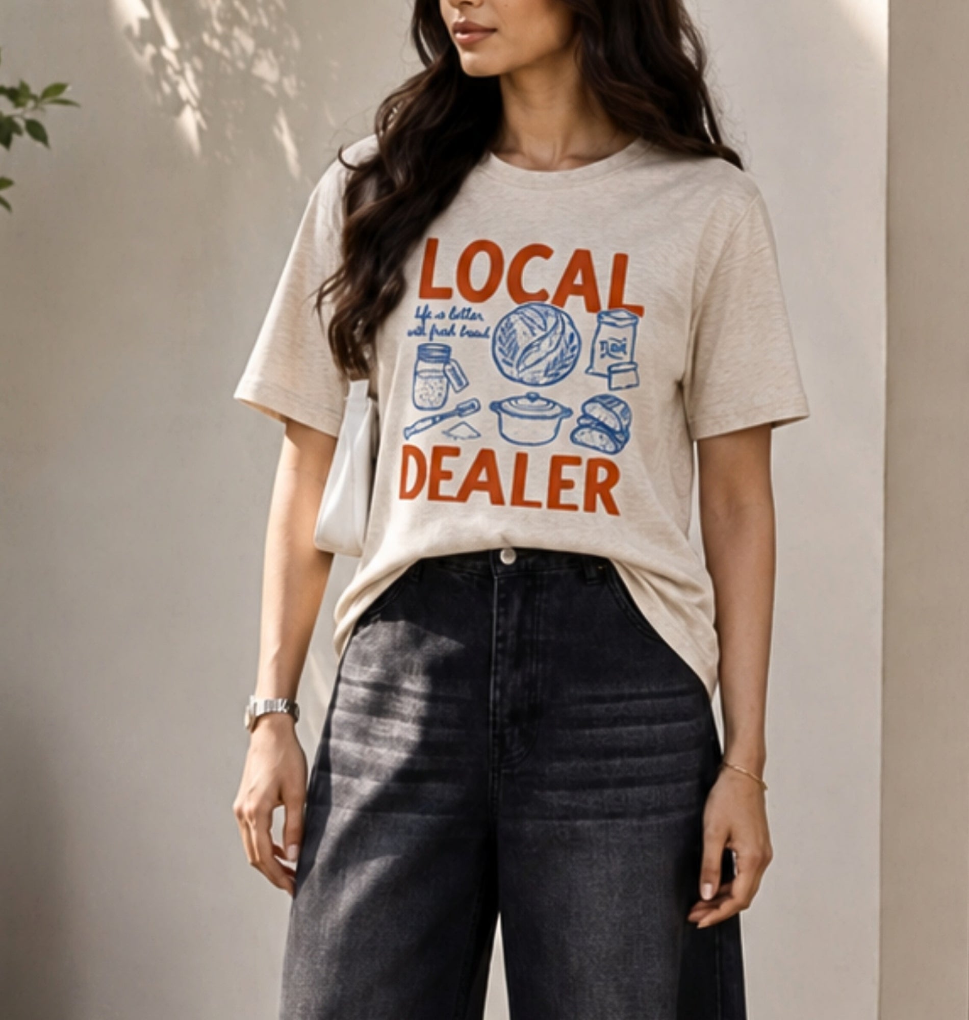 A woman wearing a beige graphic t-shirt with orange and blue text reading 'LOCAL DEALER' and dark gray wide-leg jeans, standing against a neutral wall.