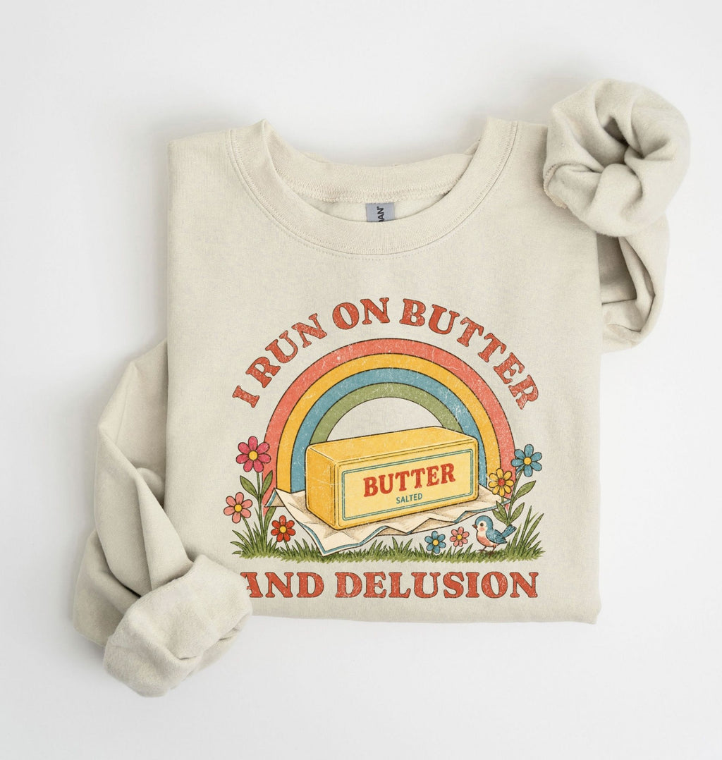 A cream-colored crewneck sweatshirt with a vintage-style graphic that reads 'I RUN ON BUTTER AND DELUSION' around a rainbow and butter block illustration. The sweatshirt is styled with one sleeve rolled up and a matching fabric scrunchie on a clean white background.