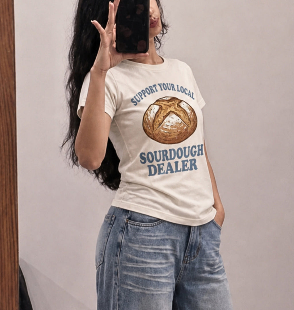 A woman takes a mirror selfie wearing a cream-colored graphic t-shirt with a sourdough bread illustration and the text 'SUPPORT YOUR LOCAL SOURDOUGH DEALER' in blue, paired with light-wash blue jeans.