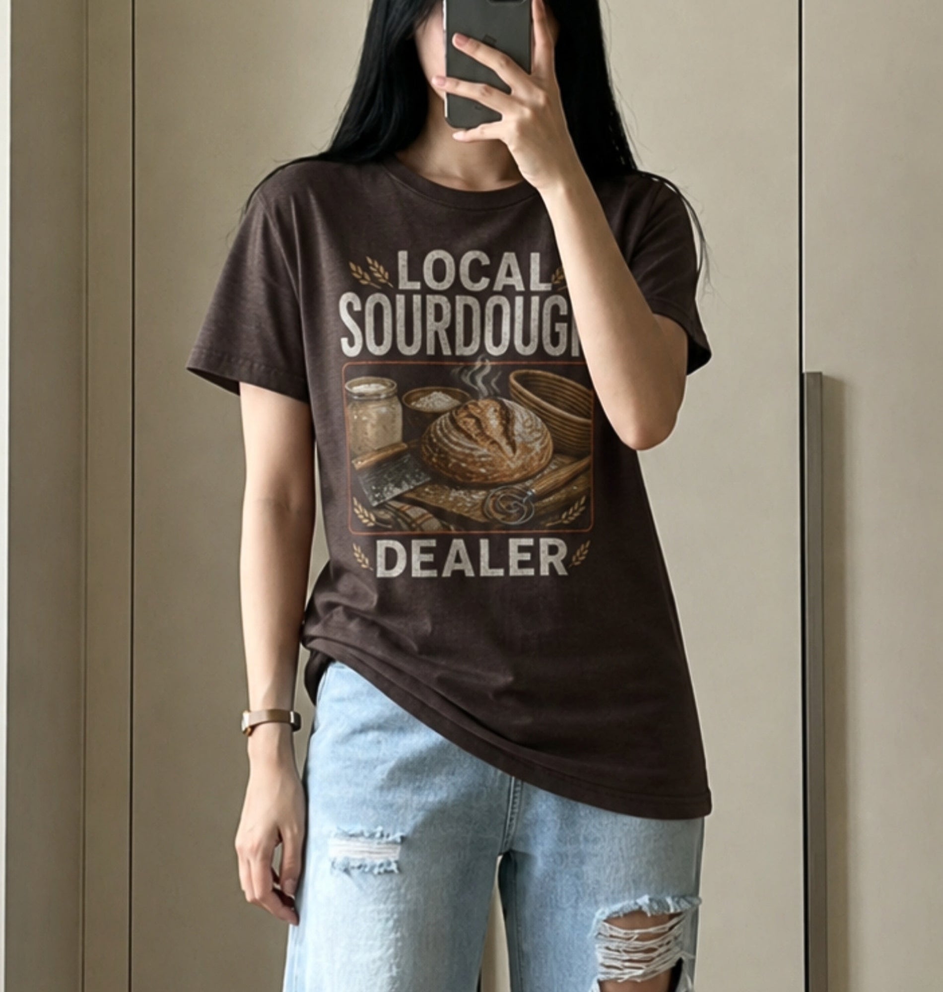 A person taking a mirror selfie wearing a dark brown graphic t-shirt with 'LOCAL SOURDOUGH DEALER' and a sourdough bread illustration, paired with light-wash distressed jeans.