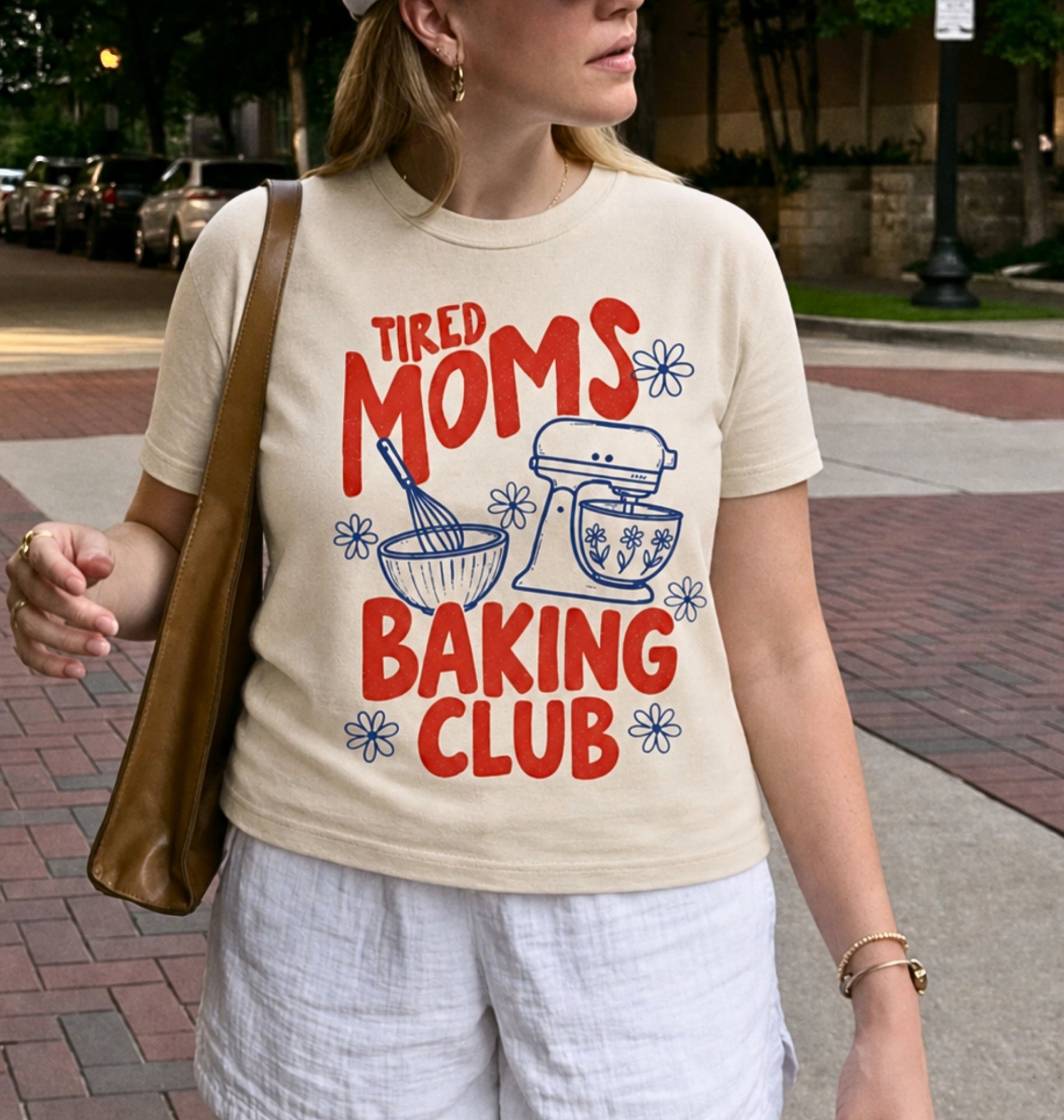 A woman wearing a beige graphic t-shirt with a red and blue 'Tired Moms Baking Club' design, paired with white shorts and a brown shoulder bag, standing on a brick sidewalk.
