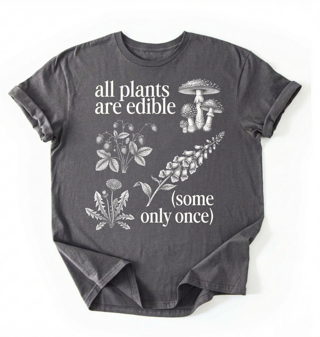 Flat lay of a Charcoal t-shirt featuring botanical illustrations and text reading “All Plants Are Edible (Some Only Once).” with mushroom graphic for plant lover gardener gift cottagecore nature shirt on a white background 