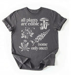 Flat lay of a Charcoal t-shirt featuring botanical illustrations and text reading “All Plants Are Edible (Some Only Once).” with mushroom graphic for plant lover gardener gift cottagecore nature shirt on a white background 