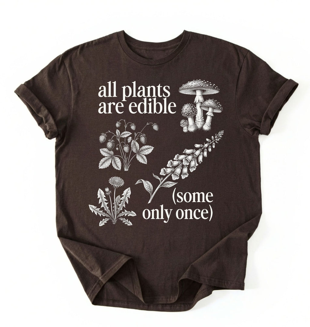 Flat lay of a brown dark chocolate shirt featuring botanical illustrations and text reading “All Plants Are Edible (Some Only Once).” with mushroom graphic for plant lover gardener gift cottagecore nature shirt on a white background 