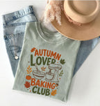 A folded sage green graphic T-shirt with the text 'AUTUMN LOVER BAKING CLUB' and autumn-themed illustrations, styled on a white surface with light blue jeans, a tan hat, and dried grass.