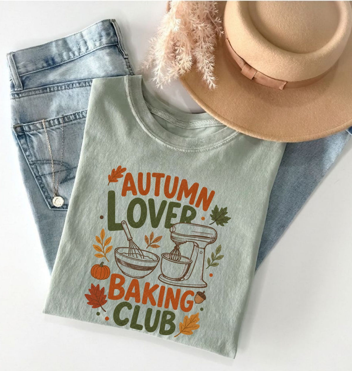 A folded sage green graphic T-shirt with the text 'AUTUMN LOVER BAKING CLUB' and autumn-themed illustrations, styled on a white surface with light blue jeans, a tan hat, and dried grass.