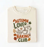 A folded cream-colored t-shirt with a colorful graphic print that reads 'AUTUMN LOVER BAKING CLUB' in autumn-themed colors, featuring a stand mixer, mixing bowl, pumpkin, and leaves.