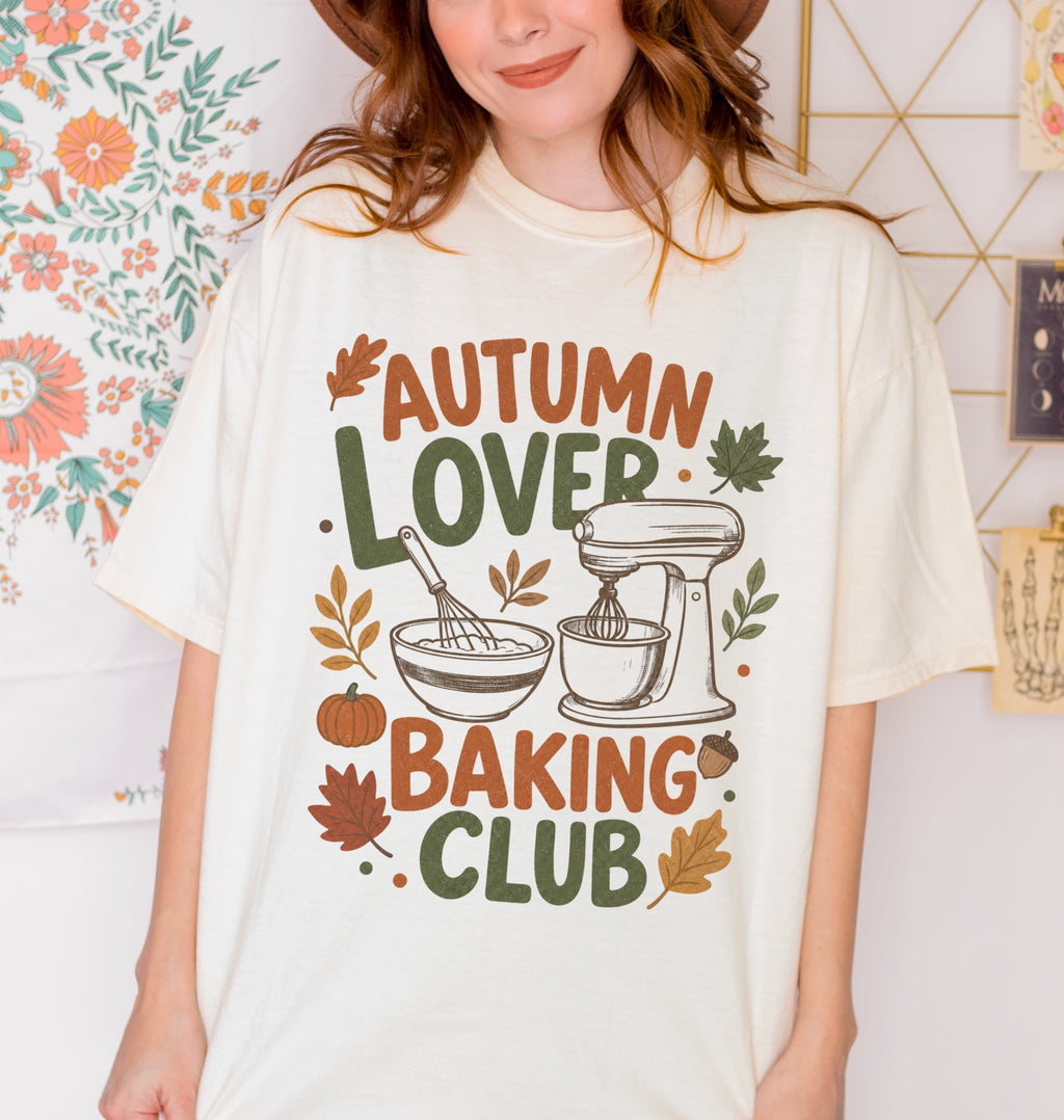 A folded cream-colored t-shirt with a colorful graphic print that reads 'AUTUMN LOVER BAKING CLUB' in autumn-themed colors, featuring a stand mixer, mixing bowl, pumpkin, and leaves.
