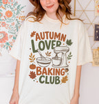 A folded cream-colored t-shirt with a colorful graphic print that reads 'AUTUMN LOVER BAKING CLUB' in autumn-themed colors, featuring a stand mixer, mixing bowl, pumpkin, and leaves.