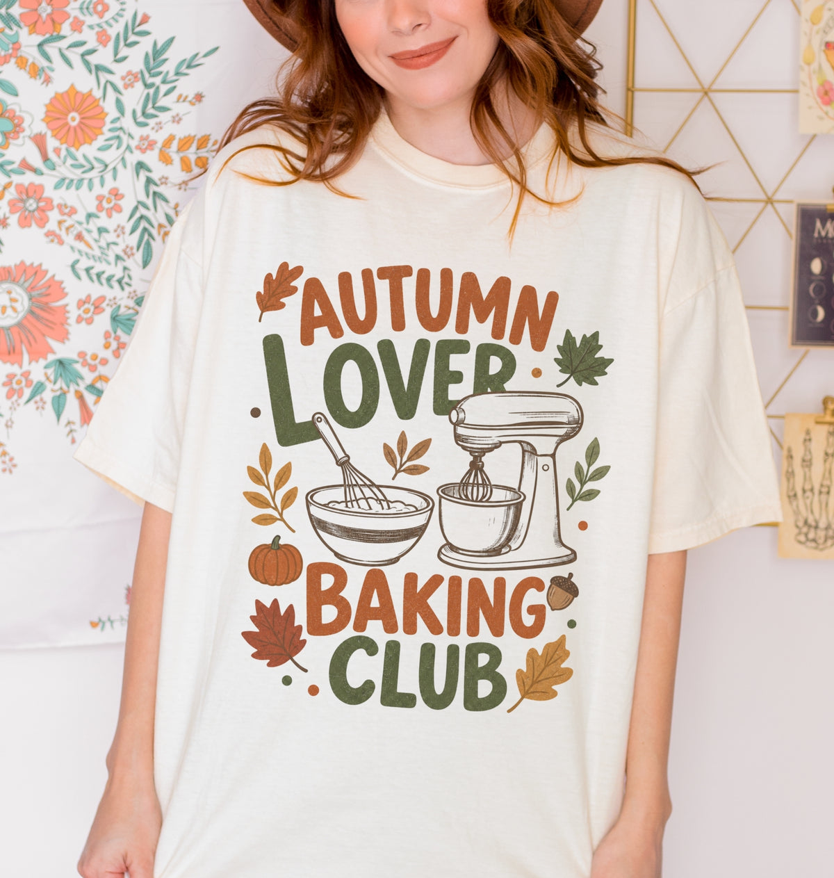 A folded cream-colored t-shirt with a colorful graphic print that reads 'AUTUMN LOVER BAKING CLUB' in autumn-themed colors, featuring a stand mixer, mixing bowl, pumpkin, and leaves.