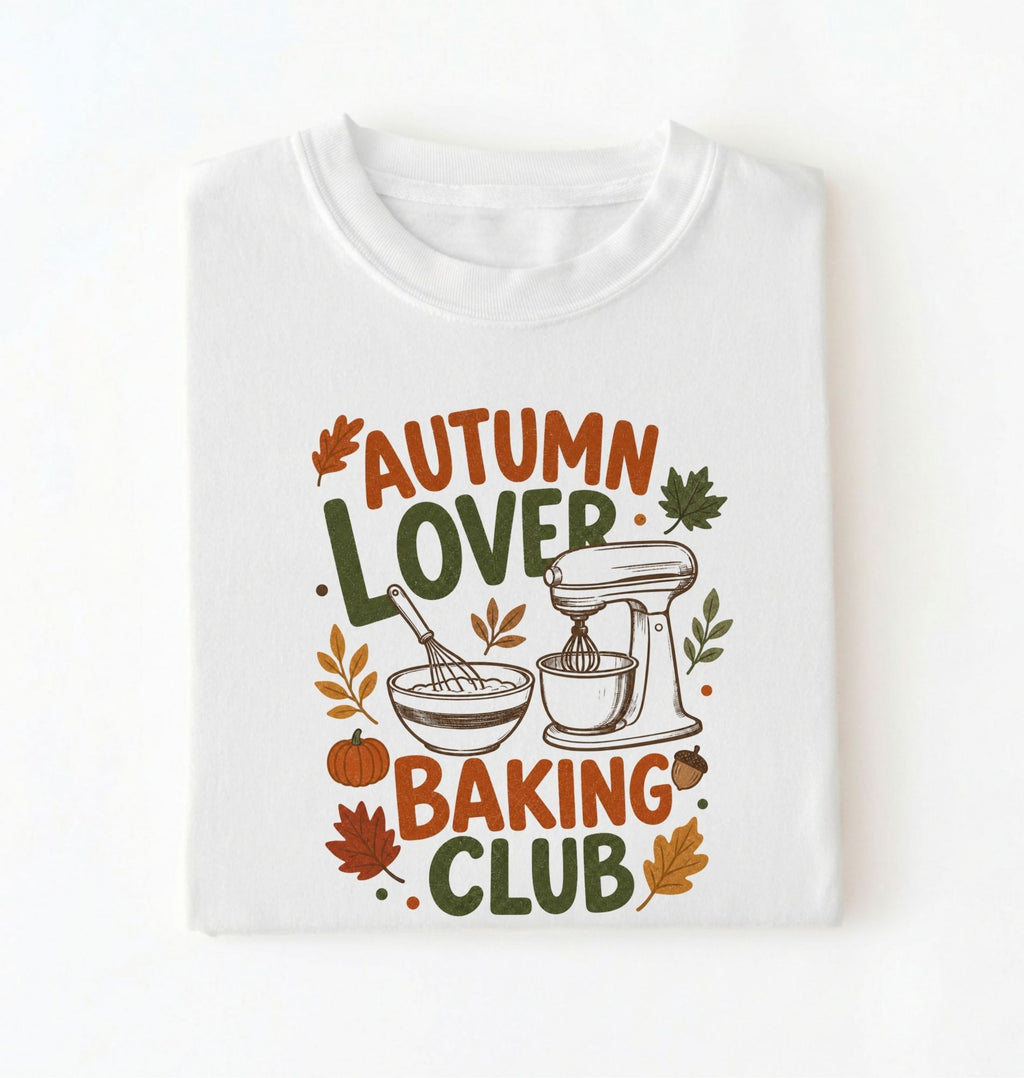 A folded white crewneck t-shirt with a colorful graphic print that reads 'AUTUMN LOVER BAKING CLUB' in autumn-themed lettering, decorated with leaves, a pumpkin, a whisk, and a stand mixer.