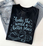 A black short-sleeve T-shirt with a light blue graphic print that reads 'bake the world a better place' with illustrations of a loaf of bread, flour sack, measuring cup, and bowl. The shirt is folded and styled on top of light-wash denim jeans against a plain white background.