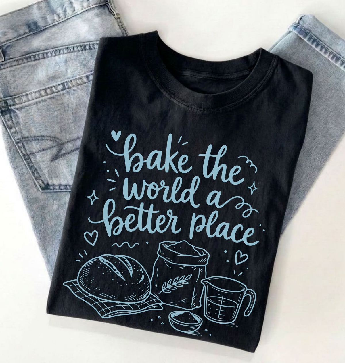 A black short-sleeve T-shirt with a light blue graphic print that reads 'bake the world a better place' with illustrations of a loaf of bread, flour sack, measuring cup, and bowl. The shirt is folded and styled on top of light-wash denim jeans against a plain white background.