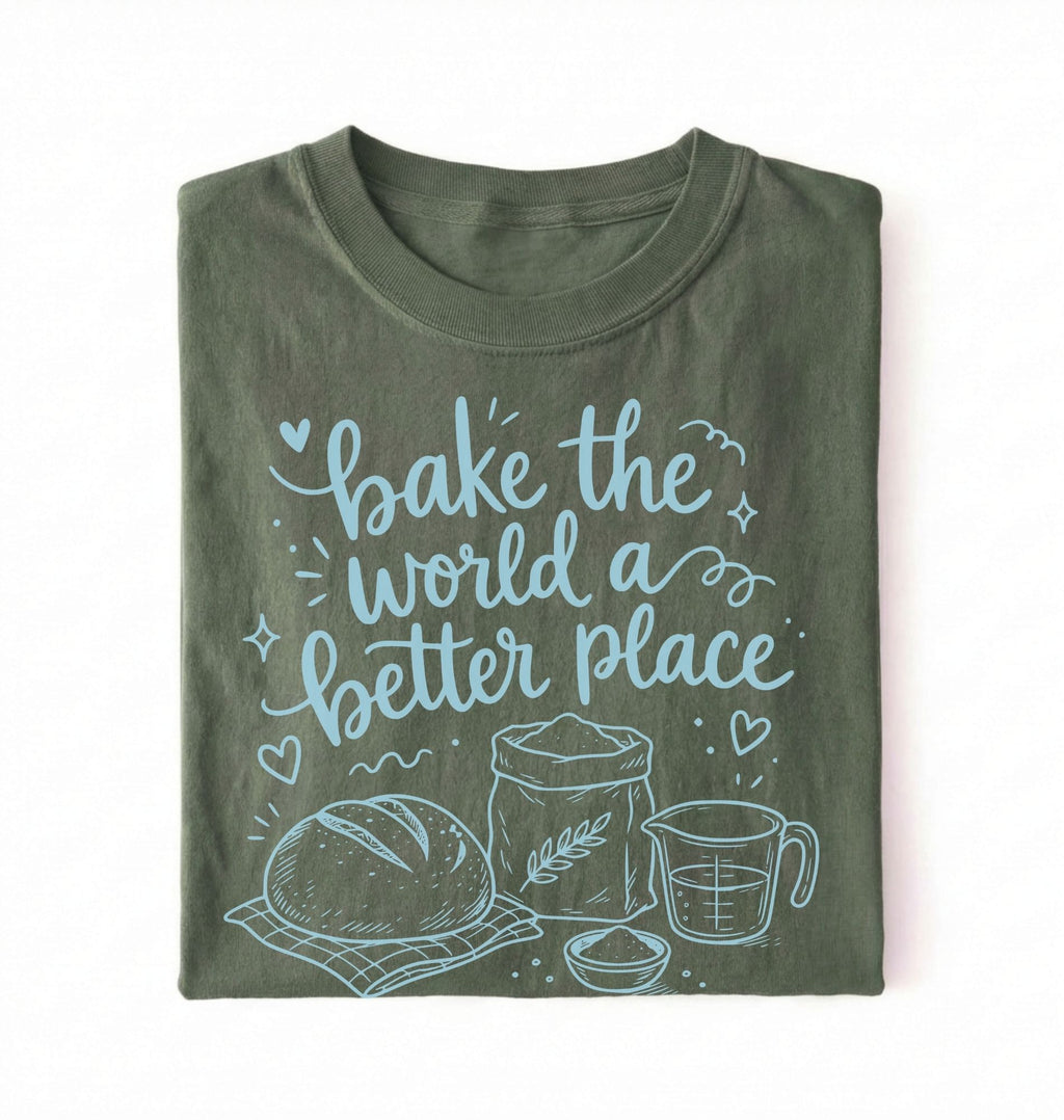 A folded dark olive green t-shirt with a light blue graphic print that reads 'bake the world a better place' accompanied by illustrations of a loaf of bread, flour, wheat, and a measuring cup, set against a plain white background.