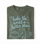 A folded dark olive green t-shirt with a light blue graphic print that reads 'bake the world a better place' accompanied by illustrations of a loaf of bread, flour, wheat, and a measuring cup, set against a plain white background.