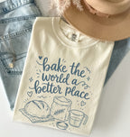 An off-white graphic t-shirt with blue lettering that reads 'bake the world a better place' and illustrations of a loaf of bread, flour sack, and measuring cup. The shirt is styled in a flat lay with light-wash denim jeans, a tan hat, and a gold necklace.