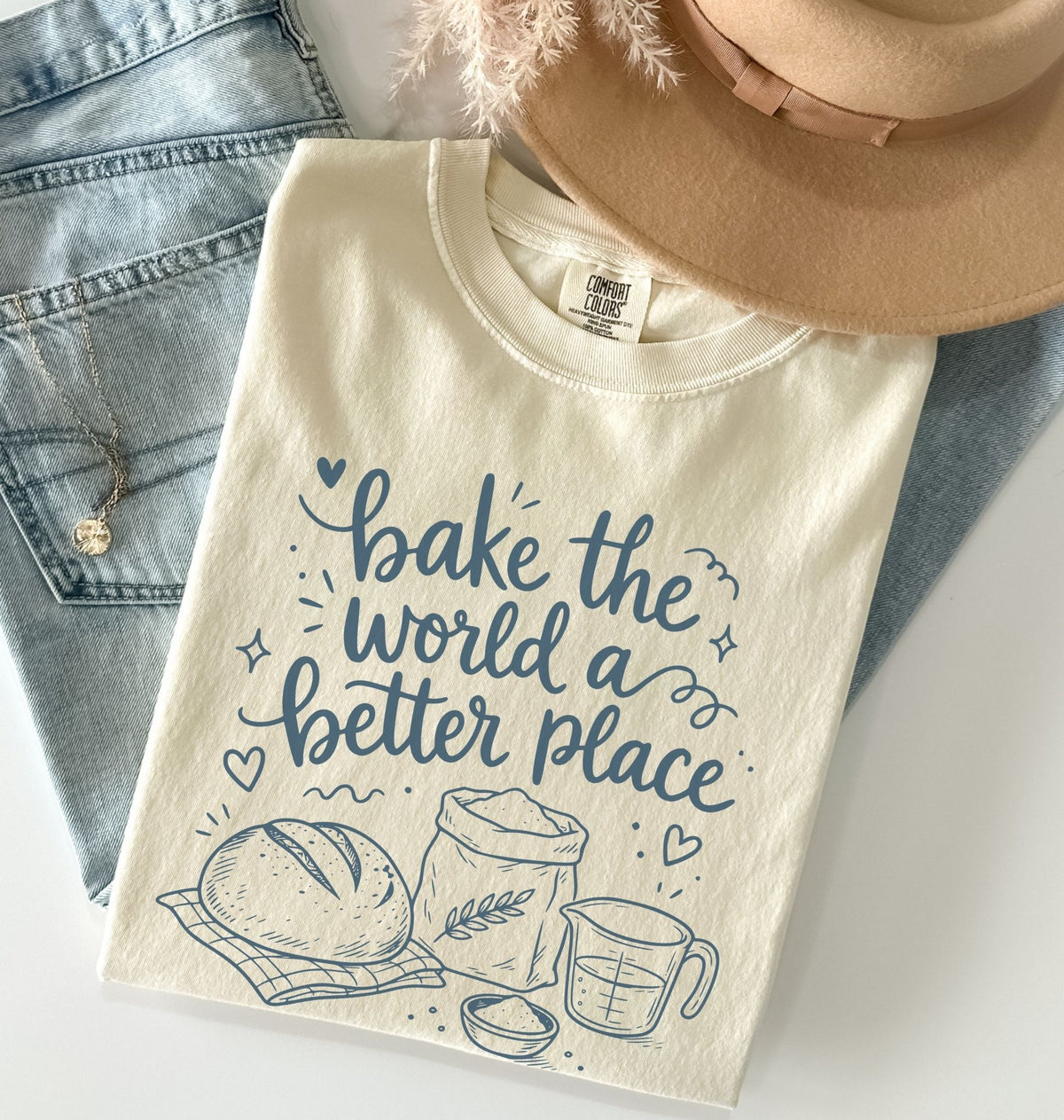 An off-white graphic t-shirt with blue lettering that reads 'bake the world a better place' and illustrations of a loaf of bread, flour sack, and measuring cup. The shirt is styled in a flat lay with light-wash denim jeans, a tan hat, and a gold necklace.