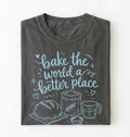 A folded dark gray t-shirt with a light blue graphic print that reads 'bake the world a better place' accompanied by illustrations of a loaf of bread, flour sack, measuring cup, and bowl, displayed on a clean white background.