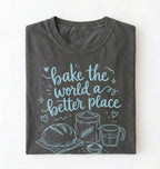 A folded dark gray t-shirt with a light blue graphic print that reads 'bake the world a better place' accompanied by illustrations of a loaf of bread, flour sack, measuring cup, and bowl, displayed on a clean white background.