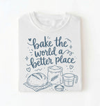 A folded white t-shirt with a navy blue graphic print that reads 'bake the world a better place' accompanied by illustrations of a loaf of bread, a bag of flour, a measuring cup, and a small bowl.
