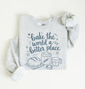 A light heather gray long-sleeve sweatshirt with a navy blue graphic print that reads 'bake the world a better place' with illustrations of a loaf of bread, flour sack, wheat stalk, measuring cup, and bowl. The sweatshirt is laid flat on a warm off-white background with one sleeve rolled up.