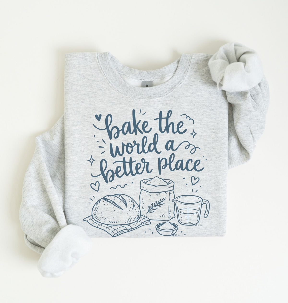 A light heather gray long-sleeve sweatshirt with a navy blue graphic print that reads 'bake the world a better place' with illustrations of a loaf of bread, flour sack, wheat stalk, measuring cup, and bowl. The sweatshirt is laid flat on a warm off-white background with one sleeve rolled up.