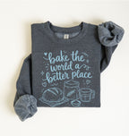 A folded dark heather gray sweatshirt with a light blue graphic print that reads 'bake the world a better place' above simple illustrations of bread, wheat, a measuring cup, and a bowl. The sleeves are styled rolled up, and the sweatshirt is photographed on a clean off-white background.