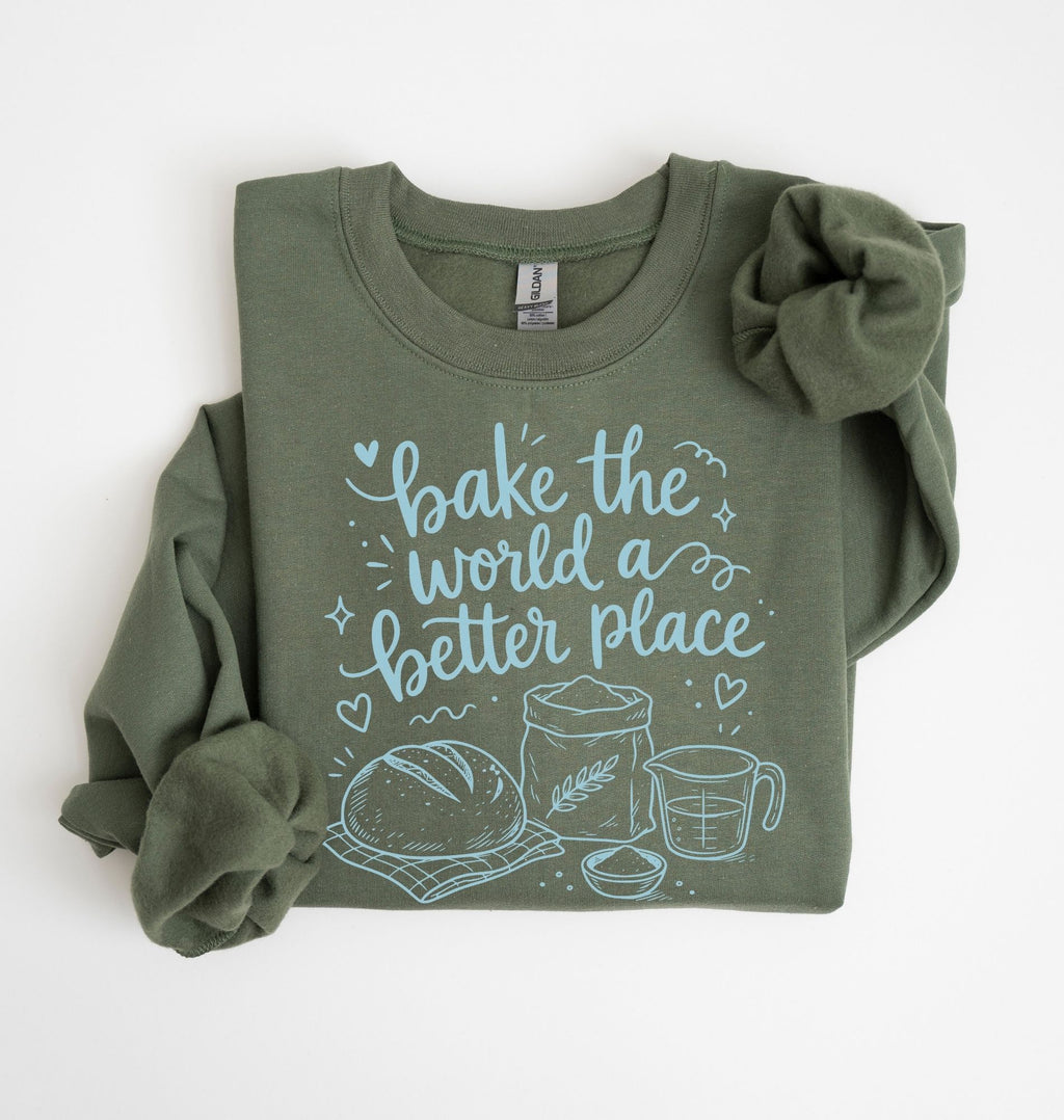 An olive green long-sleeve t-shirt with rolled sleeves, featuring a light blue graphic print that reads 'bake the world a better place' with illustrations of a loaf of bread, a wheat stalk, a measuring cup, and a bowl. The shirt is displayed flat on a clean white background.