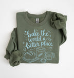 An olive green long-sleeve t-shirt with rolled sleeves, featuring a light blue graphic print that reads 'bake the world a better place' with illustrations of a loaf of bread, a wheat stalk, a measuring cup, and a bowl. The shirt is displayed flat on a clean white background.