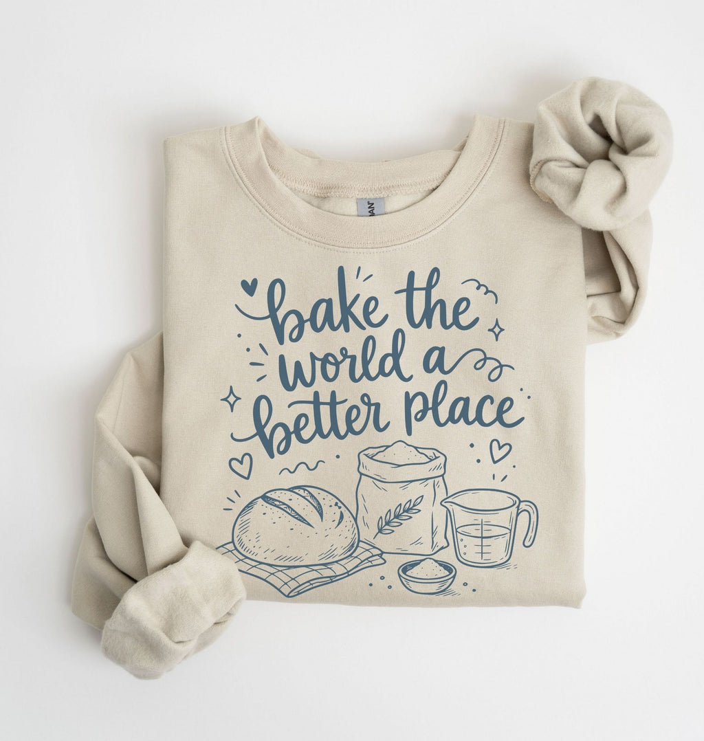 A beige crewneck sweatshirt with rolled sleeves, featuring a blue graphic print that reads 'bake the world a better place' with illustrations of a loaf of bread, a flour sack, and a measuring cup. The sweatshirt is displayed flat on a white background with a matching beige scrunchie.