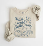 A beige crewneck sweatshirt with rolled sleeves, featuring a blue graphic print that reads 'bake the world a better place' with illustrations of a loaf of bread, a flour sack, and a measuring cup. The sweatshirt is displayed flat on a white background with a matching beige scrunchie.