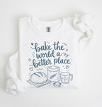 A white sweatshirt laid flat on a white background, featuring a navy blue graphic that reads 'bake the world a better place' with illustrations of a loaf of bread, a bag of flour, a measuring cup, and a small bowl.