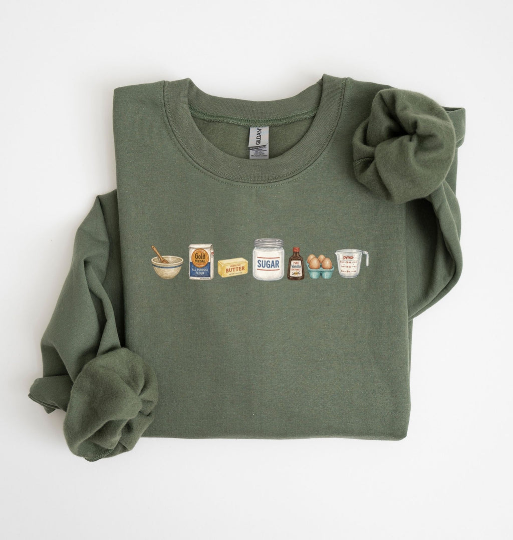 Flat lay of a green baking ingredients sweatshirt with flour butter sugar eggs vanilla and baking essentials design on a white background