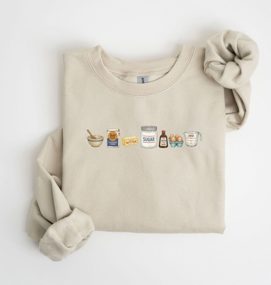 Flat lay of a sand baking ingredients sweatshirt with flour butter sugar eggs vanilla and baking essentials design on a white background