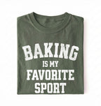 Flat lay of a green Baking Is My Favorite Sport graphic shirt on navy blue t-shirt for home bakers on a white background 