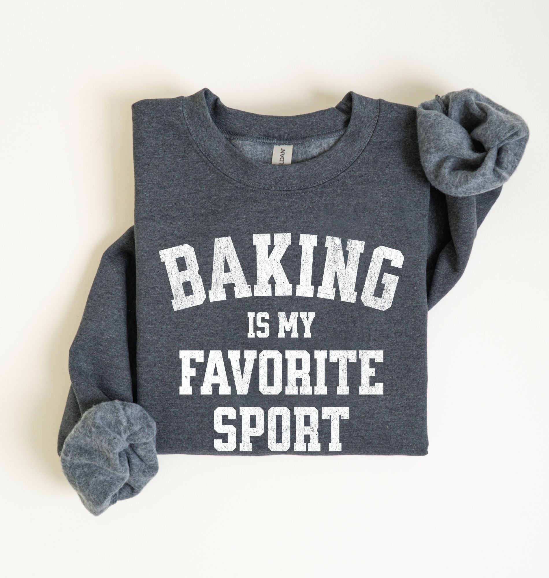 Flat lay of a dark heather Baking Is My Favorite Sport graphic sweatshirt on navy blue crewneck for home bakers on white background